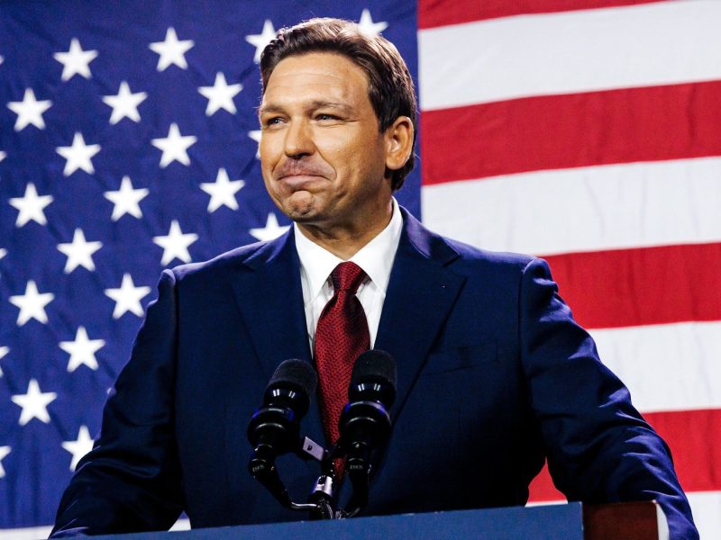 Florida Gov. Ron DeSantis wearing a dark suit and red tie standing in front of an American flag.